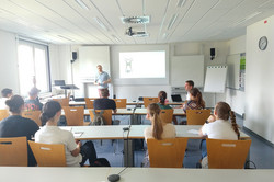 Studierende sitzen in einem Seminarraum und richten ihren Fokus auf den Dozenten, der vorne etwas erklärt.