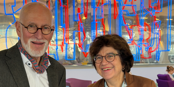 Foto: Prof. Weiler und Prof. Havenith-Newen lächeln mit Kafeetassen in der Hand in die Kamera.