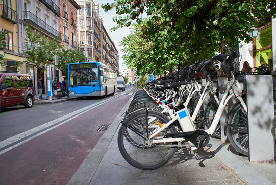 E-Bikes in der Stadt Auf einer Straße stehen rechts mehrere E-Bikes in einer Reihe und links hält ein blauer Bus, die Bäume sind grün im Hintergrund.
