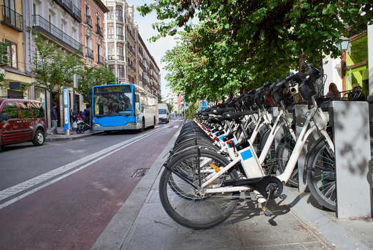 Auf einer Straße stehen rechts mehrere E-Bikes in einer Reihe und links hält ein blauer Bus, die Bäume sind grün im Hintergrund.