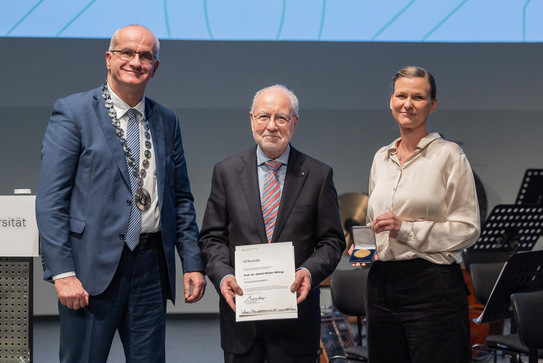 Das Bild zeigt Bayer, Müller-Böling, Flatten, welche nebeneinanderstehend in die Kamera lächeln. Müller-Böling hält eine Urkunde für eine weitere Universitätsmedaille in seinen Händen.