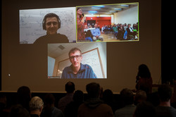 Auf einer großen Projektionsleinwand ist ein Zoom-Videocall mit zwei jungen Männern und einem Raum voller Zuhörer*innen zu sehen.