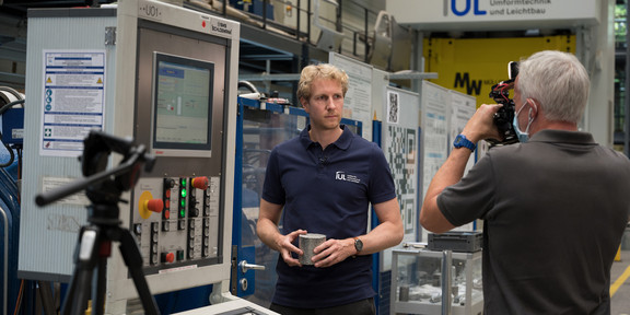 André Schulze IUL Alurecycle Andre Schulze steht vor einem Gerät im Institut für Umformtechnik und Leichtbau der TU Dortmund, während ein Mann seine Kamera auf ihn richtet.
