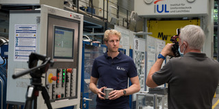 Andre Schulze steht vor einem Gerät im Institut für Umformtechnik und Leichtbau der TU Dortmund, während ein Mann seine Kamera auf ihn richtet. 