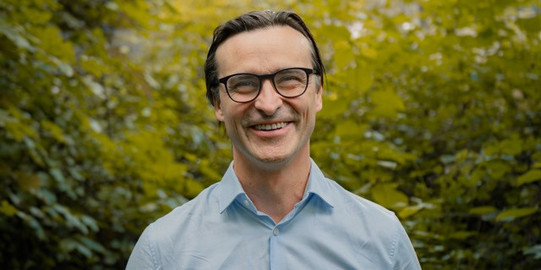 Porträtfoto von Benedikt Sabaß: Prof. Sabaß trägt ein hellblaues Hemd und lacht in die Kamera. Im Hintergrund sind Büsche.