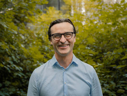 Porträtfoto von Benedikt Sabaß: Prof. Sabaß trägt ein hellblaues Hemd und lacht in die Kamera. Im Hintergrund sind Büsche.