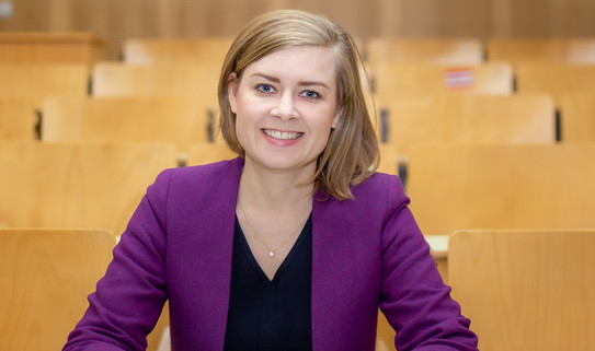 Eine Frau (Dr. Carolin Funke) sitz in der Tischreihe eines Hörsaals und posiert für das Foto.