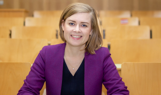 Eine Frau (Dr. Carolin Funke) sitz in der Tischreihe eines Hörsaals und posiert für das Foto.