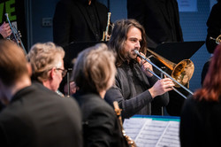 Photo: A trumpet player with long hair plays his instrument. Other musicians in dark clothing sit around him.