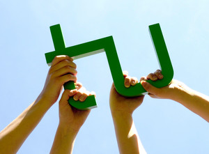 Vier Hände halten ein hölzernes, grün lackiertes TU-Logo in den blauen Himmel