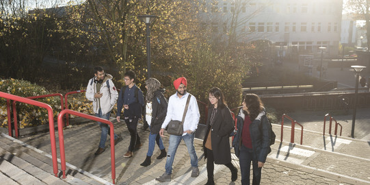 Sechs Student*innen laufen zusammen die Treppen am Campus hoch und unterhalten sich