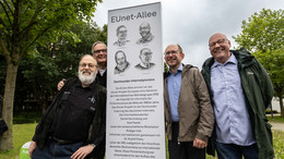 Juli vier Männer stehen draußen vor einer weißen Stele, auf der ihre Gesichter (in einem Fall das des Vaters) gezeigt sind
