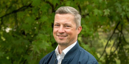 Porträtfoto von Prof. Matthias Kortmann: Prof. Kortmann trägt ein helles Hemd und eine dunkle Jacke und lächelt in die Kamera.