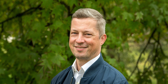Portrait photo of Prof. Matthias Kortmann: Prof. Kortmann is wearing a light-colored shirt and a dark jacket and smiling at the camera.