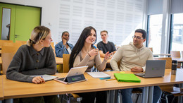 Oktober Fünf Studierende sitzen in einem Seminarraum mit grüner Tür. Im Vordergrund sitzen drei Studierende, vor denen iPads und Laptops stehen.