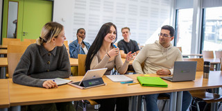 Fünf Studierende sitzen in einem Seminarraum mit grüner Tür. Im Vordergrund sitzen drei Studierende, vor denen iPads und Laptops stehen.