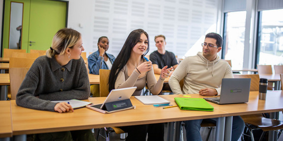 Fünf Studierende sitzen in einem Seminarraum mit grüner Tür. Im Vordergrund sitzen drei Studierende, vor denen iPads und Laptops stehen.