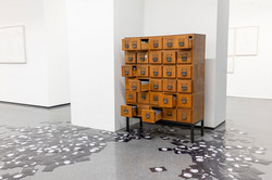 Ausstellung „Laufende Beobachtungen“ In einer Ausstellung steht ein brauner Schrank mit Schubladen an einer Wand. Mehrere Schubladen sind geöffnet.