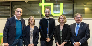 Five people in dark jackets and blazers in front of a TU logo.