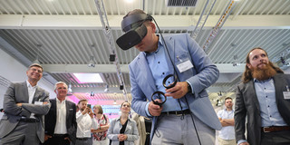 A man in a light blue jacket is trying out VR glasses. Around him, other people stand in a circle.