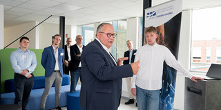 Im Vordergrund zu sehen ist Josef Hovenjürgen und ein Mitarbeiter der UA Ruhr. Im Hintergrund stehen weitere Mitarbeiter der UA Ruhr und der Rektor der TU Dortmund. Sie stehen in einem Büro vor einer blauen Couch und einer grünen Trennwand.