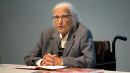 Horst Selbiger, ein älterer Mann mit grauem Zopf und Brille, sitzt im Rollstuhl an einem Tisch und spricht. vor ihm liegen Bücher und ein aufgeschlagener Ordner mit Text.