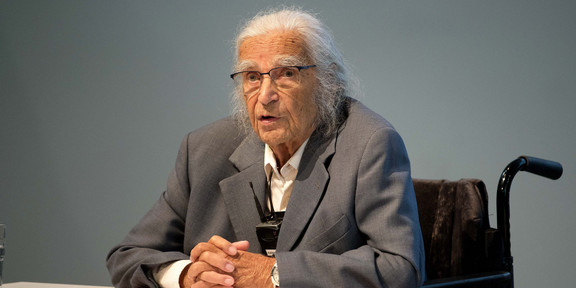 Horst Selbiger, ein älterer Mann mit grauem Zopf und Brille, sitzt im Rollstuhl an einem Tisch und spricht. vor ihm liegen Bücher und ein aufgeschlagener Ordner mit Text.