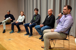 Podiumsdiskussion. Auf der Bühne sitzen fünf Personen und ganz rechts spricht ein Mann in das Mikrofon.