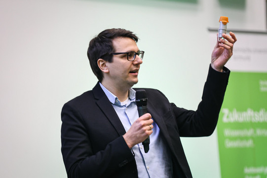 Dr. Raphael Krampe, Projektleiter Entwicklung Kombinationsprodukte bei Boehringer Ingelheim microParts, spricht ins Mikrofon, in der linken Hand hält er einen Tascheninhalator hoch