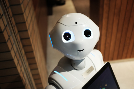 Weißer humanoider Roboter mit großen leuchtenden Augen steht in einem Innenraum neben einer Ziegelwand und hält ein Tablet in den Händen.