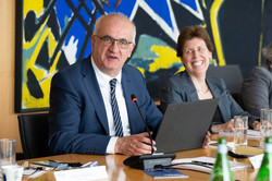 Im Zentrum des Bildes ist Prof. Bayer sitzend hinter einem Tisch und vor einen Laptop zu sehen. Rechts neben ihm sitzt eine lachende Frau.