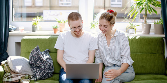 Eine junge Frau und ein junger Mann sitzen auf einem Sofa und schauen gemeinsam auf einen Laptop.