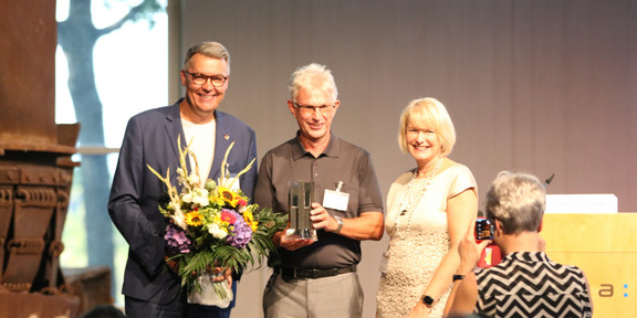 Two men and a woman stand on a stage for an award ceremony.