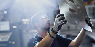 Foto: Ein Mann mit Schutzbrille und Arbeitshandschuhen hält eine silberne Metallplatte mit Ausstanzungen hoch und schaut sie prüfend an. 