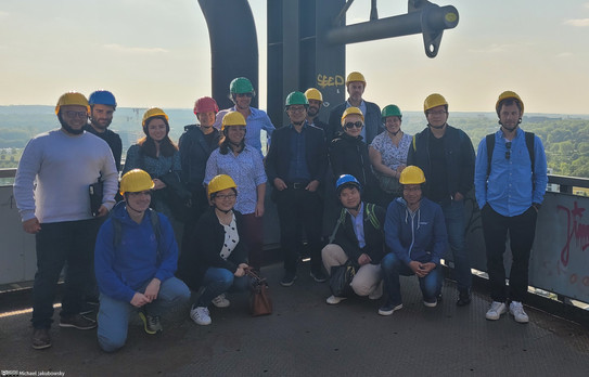 Kick-Off Healing Bat Eine Gruppe Menschen steht auf der Plattform eines hohen Gebäudes, alle tragen Schutzhelme. Im Hintergrund sind Hügel und Bäume zu sehen.