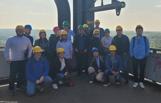 A group of people are standing on the platform of a tall building, all wearing hard hats. Hills and trees can be seen in the background.