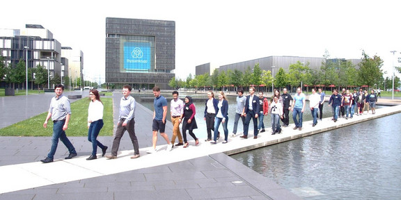 A large group of young adults walk across a footbridge over an expanse of water in front of the thyssenkrupp headquarters building