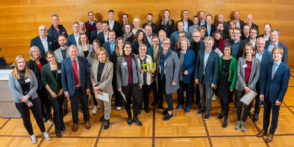 Zu sehen ist die Gruppe an Stifter*innen bei der Stipendienfeier. Eine Person in de Mitte hält das grüne TU-Logo in der Hand. 