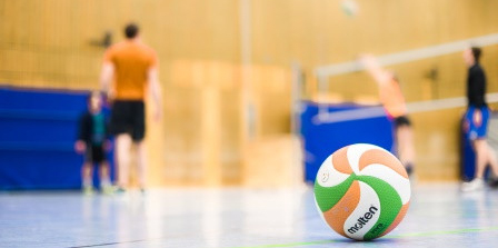 In einer Sporthalle liegt ein Volleyball, im Hintergrund sind Sportlerinnen und und Sportler