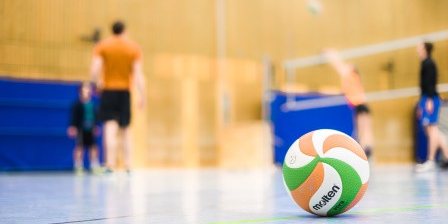 In einer Sporthalle liegt ein Volleyball, im Hintergrund sind Sportlerinnen und und Sportler