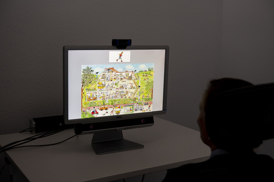 Eine Frau sitzt vor einem hellen Computer-Bildschirm, der ein Wimmelbild zeigt. Auf dem Computer ist eine Webcam angebracht. 
