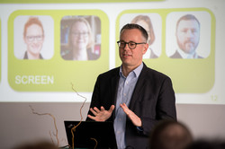 Das Bild zeigt eine männlich gelesene Person im Anzug hinter einem Rednerpult. Im Hintergrund ist eine Leinwand mit zwei grünen Kacheln zu sehen, in denen sich wiederum jeweils zwei Porträtfotos von Personen befinden.