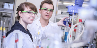 Eine auszubildende Chemielaborantin und ein auszubildender Chemielaborant stehen mit Schutzkleidung in einem Labor.