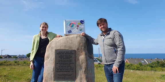Eine Frau und ein Mann stehen neben einem hohen Stein, auf dem ein silberner Koffer mit Stickern steht. Im Hintergrund sieht man Häuser und das Meer.