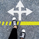 You can see the legs and the feet of a person standing on asphalt ground. In front of the person's feet there are three white arrows in the ground, pointing forward, to the right and to the left.