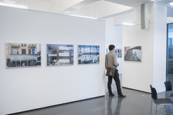 Ein Mann steht zwischen Ausstellungswänden und sieht sich eine Fotografie an. Sie zeigt eine Spiegelung in einer Autotür. Auf der Wand links sind Fassadenansichten zu sehen.