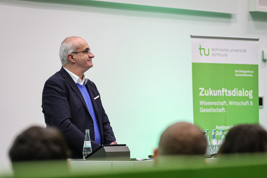 ein Mann steht vor Publikum, rechts im Hintergrund steht ein Banner ein grün-weißes Banner mit der Aufschrift "Zukunftsdialog"