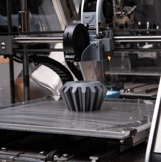Ein 3D-Drucker fertigt ein gezacktes, zahnradähnliches Bauteil aus dunklem Kunststoff auf einer Druckplatte an.