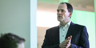 Das Bild zeigt Dr. Hendrik Neumann in einem schwarzen Jaquette mit weißem Hemd. Er hält seine Hände flach zusammengedrückt auf höhe seiner Brust. Unten links im Bild ist unscharf der Kopf einer Person zu erkennen.