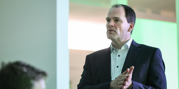 Das Bild zeigt Dr. Hendrik Neumann in einem schwarzen Jaquette mit weißem Hemd. Er hält seine Hände flach zusammengedrückt auf höhe seiner Brust. Unten links im Bild ist unscharf der Kopf einer Person zu erkennen.